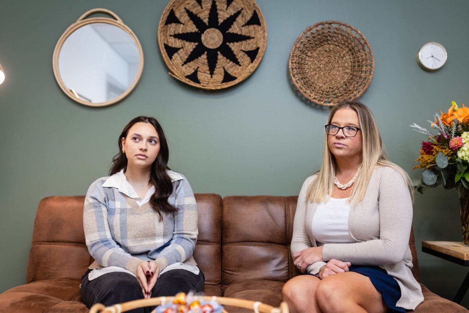 Two women sitting on a couch in front of wall hangings.